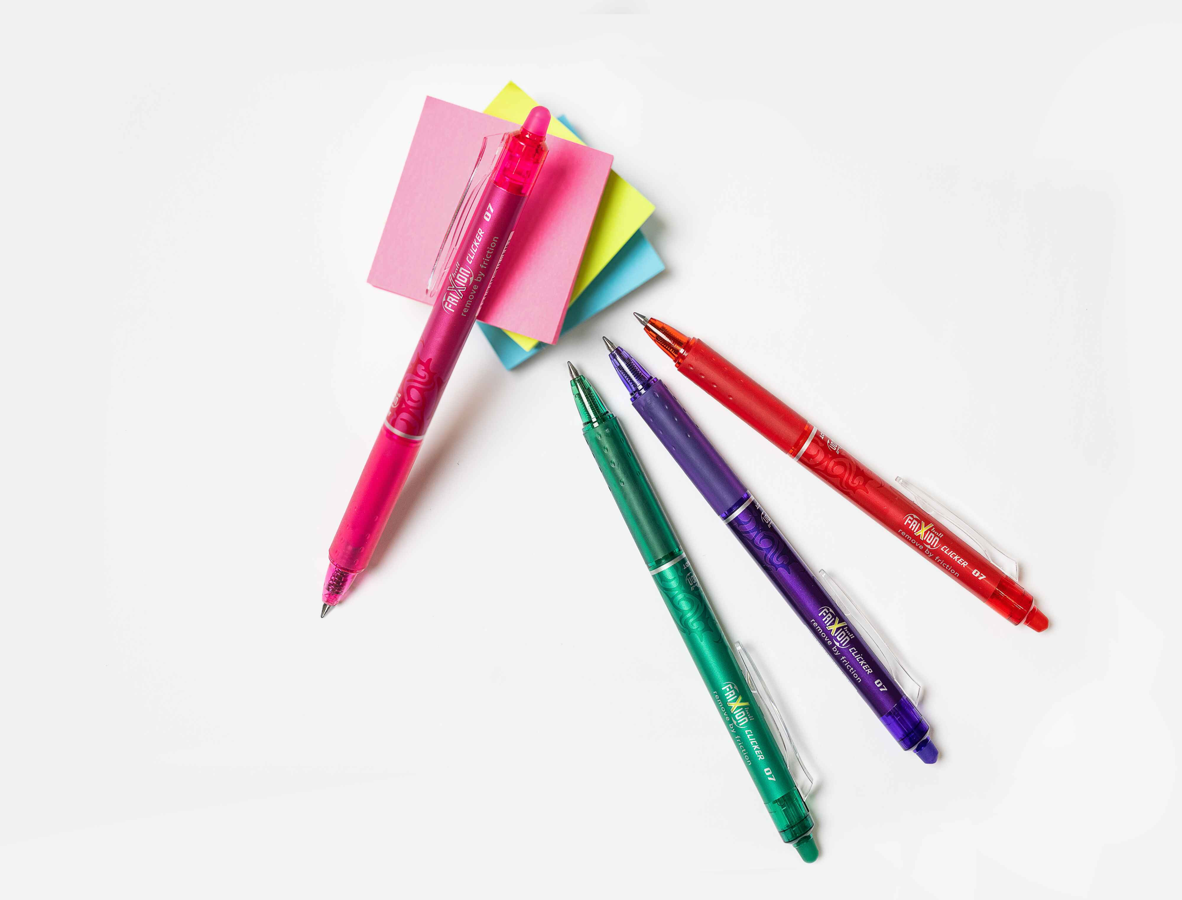 A selection of different coloured refillable FriXion Clicker pens and a post-it pad arranged on a white surface.